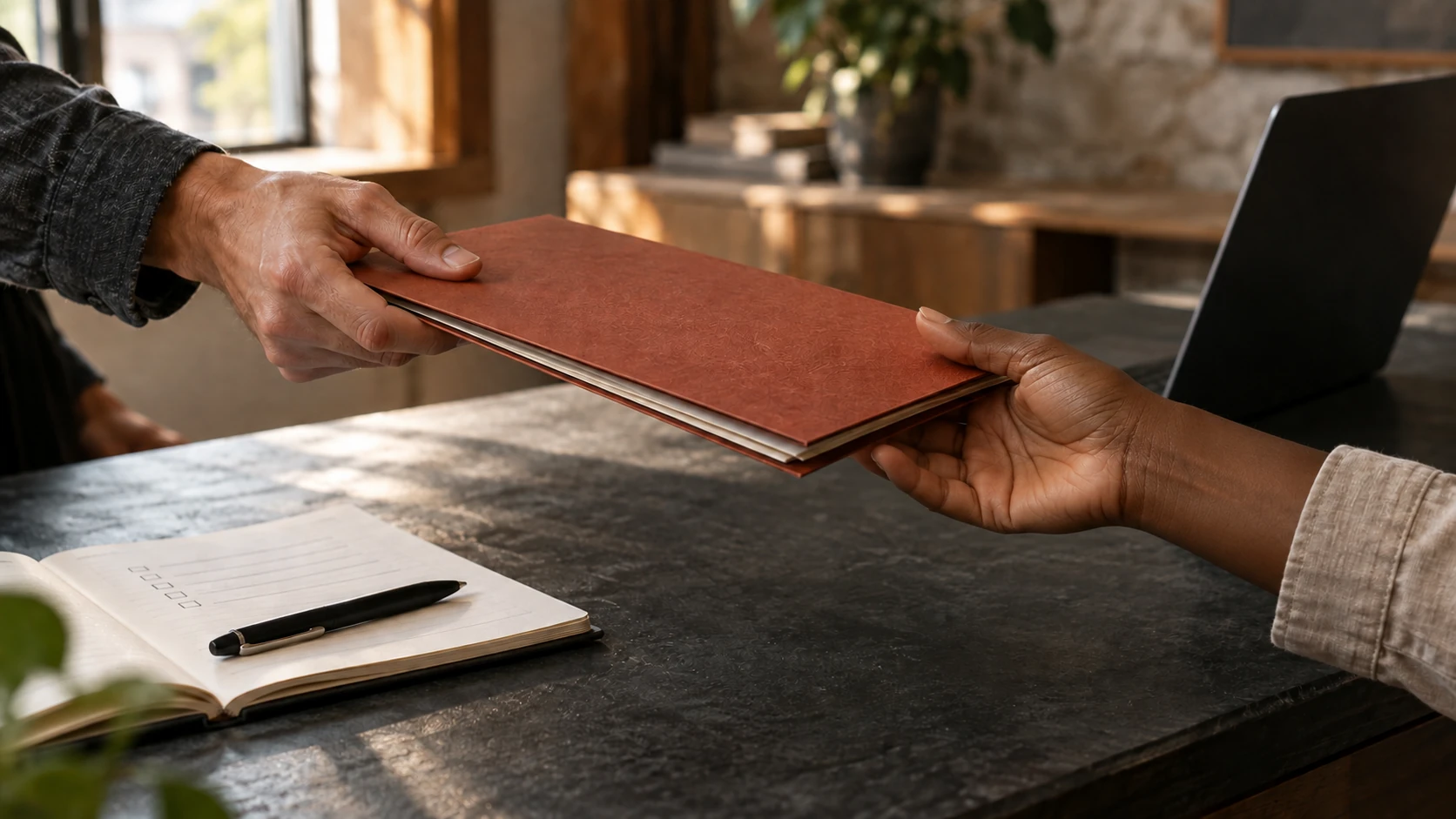 Two people pass a rust-colored document folder across a worktable beside a notebook.